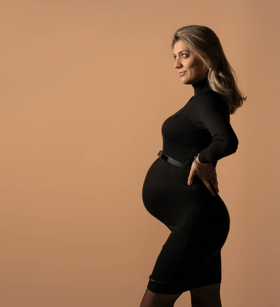 Mujer embarazada en vestido negro apretado posando en el estudio sobre un fondo beige. - Foto, imagen