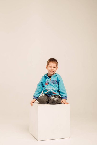 Een kleine jongen in een blauwe blouse zit op een witte kubus in een heldere kamer - Foto, afbeelding