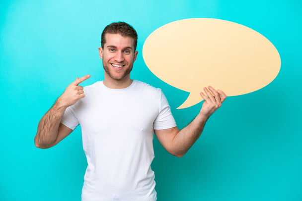 Young caucasian man isolated on blue background holding an empty speech bubble and pointing it - Photo, Image