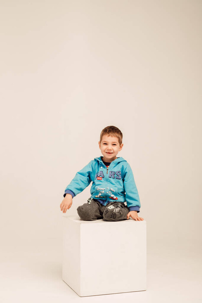 Un niño pequeño en una blusa azul se sienta en un cubo blanco en una habitación luminosa - Foto, Imagen