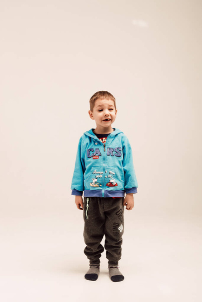 Un niño pequeño con una chaqueta azul se levanta sobre un fondo blanco y sonríe - Foto, Imagen