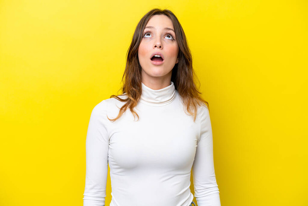 Mujer joven caucásica aislada sobre fondo amarillo mirando hacia arriba y con expresión sorprendida - Foto, Imagen