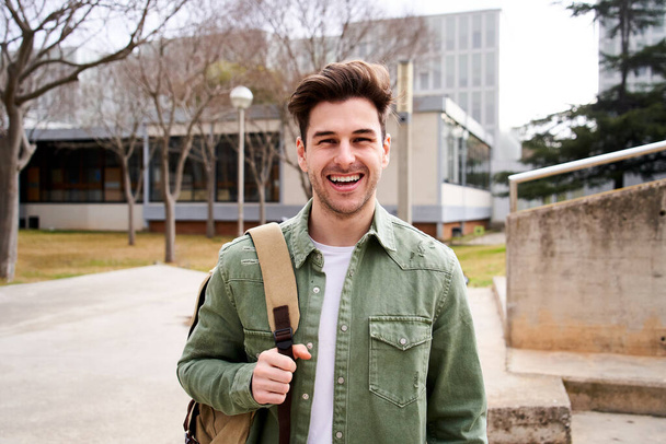 Nahaufnahme eines fröhlichen kaukasischen Teenagers, der lächelnd in die Kamera blickt. Lustiges Porträt eines jungen Studenten mit seinem Rucksack an der Campus-Universität. Kerl lacht an der High School. - Foto, Bild