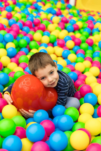 A little boy sits in bright balls and holds in his hands a red ball  - Фото, изображение