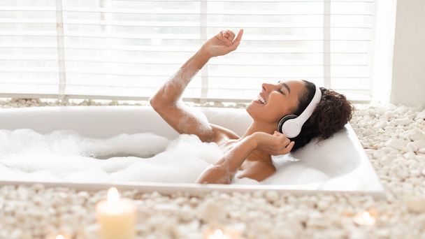 Позитивная юная леди в наушниках принимает пенную ванну, слушает музыку и поет любимую песню дома, баннер - Фото, изображение