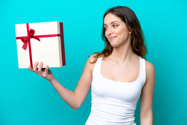 Молода біла жінка тримає подарунок ізольовано на синьому фоні з щасливим виразом
 - Фото, зображення