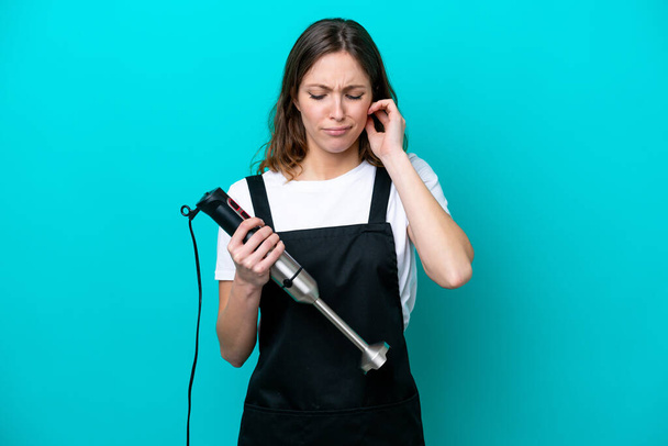 Mujer joven cocina caucásica usando licuadora de mano aislada sobre fondo azul frustrado y cubriendo oídos - Foto, imagen