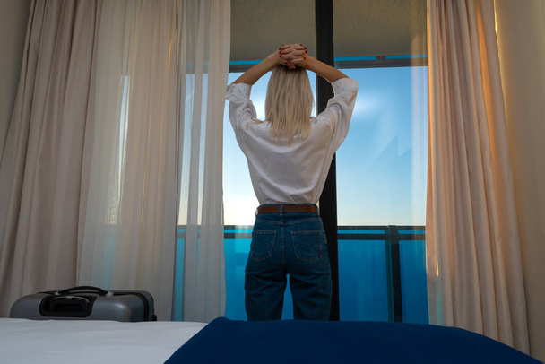 白いシャツを着た若い女性と青いジーンズは、ホテルの部屋で彼女の頭の上に手で窓に立っています。旅行と夏休みの概念. - 写真・画像