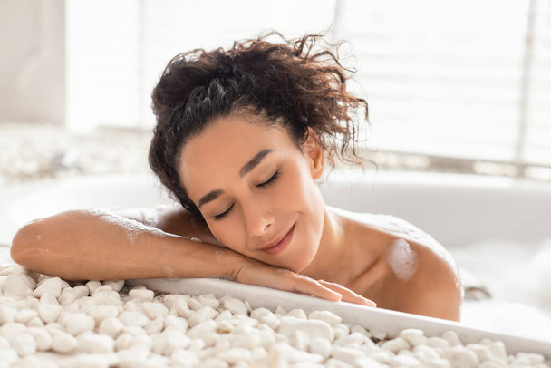 Beautiful young woman lying in bubble bath with closed eyes at home. Beauty and wellness concept - Photo, Image