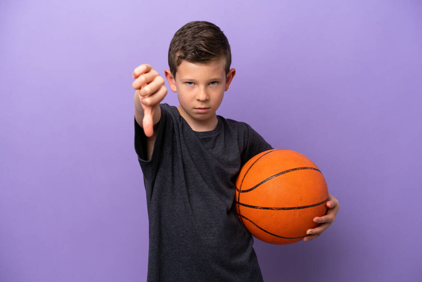 小さな男の子は、負の式で親指を示す紫色の背景に隔離されたバスケットボールをプレイ - 写真・画像