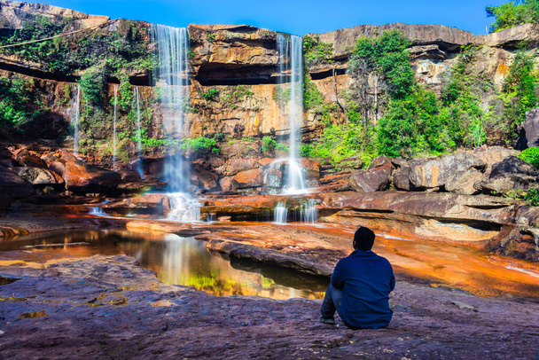 odizolowany młody człowiek siedzący w pobliżu wodospadu spadającego z góry z odbiciem i błękitnym niebem - Zdjęcie, obraz