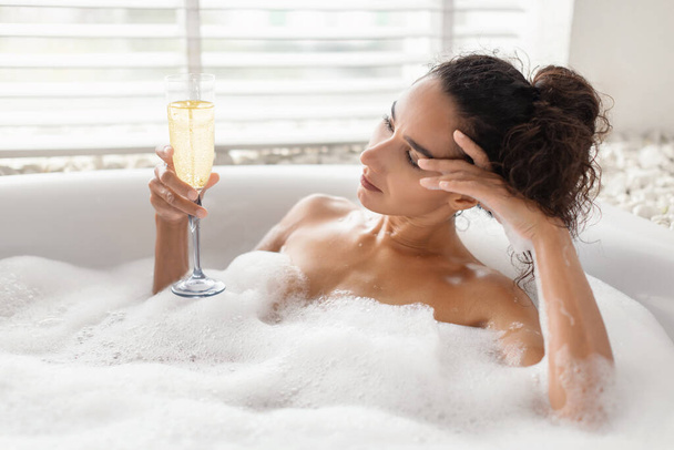 Jovem infeliz deitada em banho de espuma com copo de champanhe em casa, espaço de cópia - Foto, Imagem