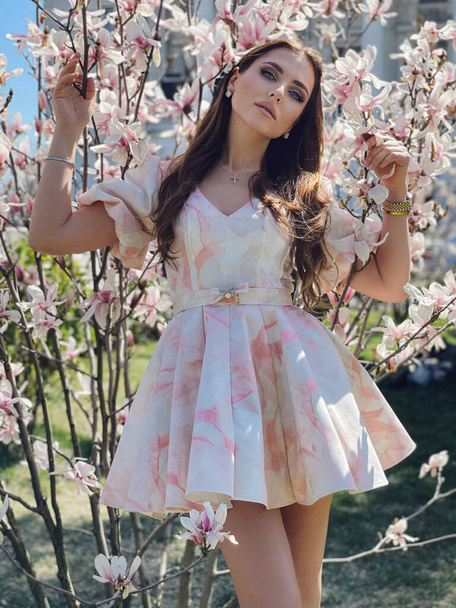 fashion outdoor photo of beautiful woman with dark hair in elegant dress posing in flowering spring park with magnolias - Фото, зображення