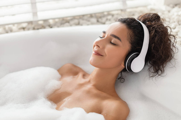 Mujer joven despreocupada escuchando música tranquila, disfrutando de la meditación mientras está acostada en el baño de burbujas en casa, espacio para copiar - Foto, imagen