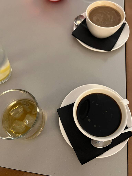 Interior view of coffee drinks in white cups sat on black serviette napkin with saucers and spoons, vegan one black other oat milk, glass of brandy spirit with ice, jug lemon water flat lay on table - Zdjęcie, obraz