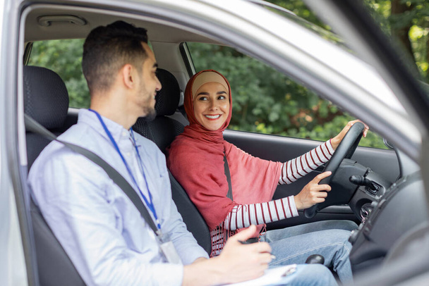 Sonriente milenaria de Oriente Medio hembra en hijab en el volante de conducción de coches tomando la prueba de conducción con el profesor - Foto, imagen