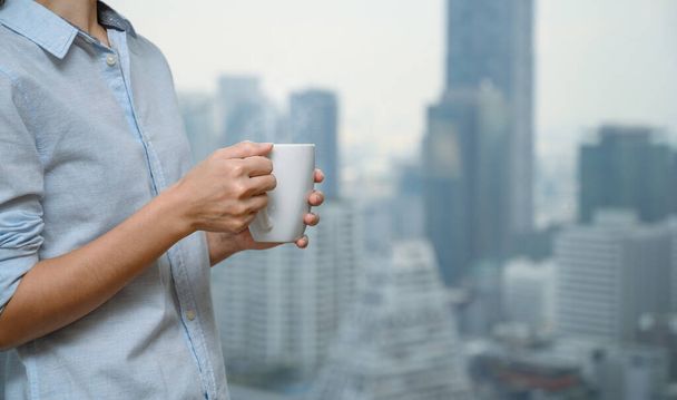 Primo piano mani di donna in possesso di una tazza di caffè o tè bianco dalla finestra con sfocato di vista sulla città alto edificio sullo sfondo. Copia spazio - Foto, immagini