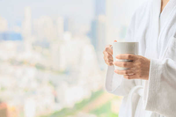 Vista laterale della donna in accappatoio in piedi vicino alla finestra mentre tiene in mano una tazza di caffè bianco e godersi una splendida mattina in hotel, staccandosi dal lavoro - Foto, immagini