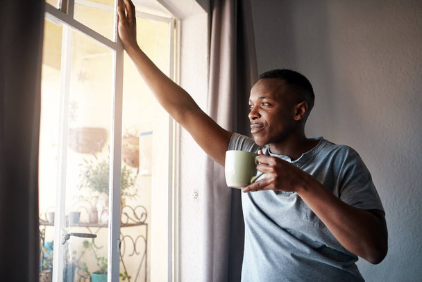 I wonder what I should do today. Cropped shot of a handsome young man standing and looking contemplative in his bedroom while enjoying a cup of coffee. - Fotografie, Obrázek