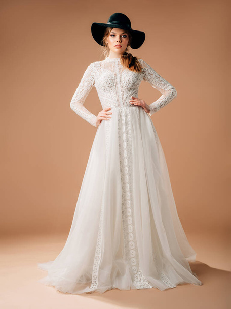 Rustic wedding dress. Elegant bridal gown with tender cotton lace, long transparent sleeves, lush tulle skirt. Beautiful young blonde lady bride with black cowboy hat supply. Studio shot. - 写真・画像