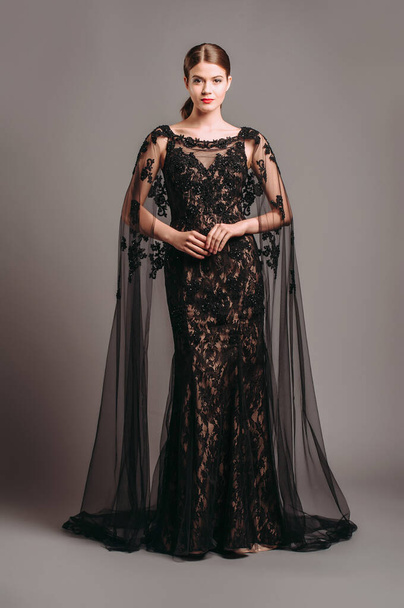 Luxury lace evening gown in black with transparent flutter cape. Embroidered maxi dress with a train, modern fashion. Fishtail fitted dress. Brunette model posing in studio. Total black female look.  - Photo, Image