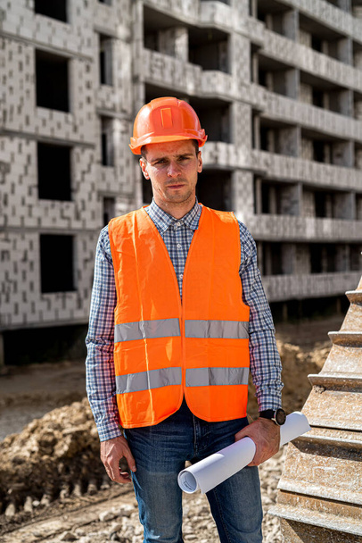 een sterke man in uniform en helm staat onder een onafgewerkt huis op een bouwplaats. Handen van een bouwvakker met een plan. Bouwconcept - Foto, afbeelding
