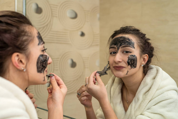 mooi jong meisje maakt een zwart masker voor acne op het gezicht. Acne masker concept - Foto, afbeelding