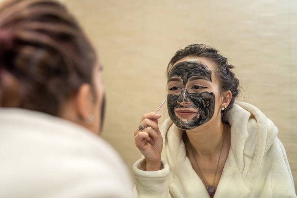 la fille dans la salle de bain se examine dans le miroir et corrige le masque de beauté nouvellement appliqué sur son visage. Concept de masque de beauté - Photo, image