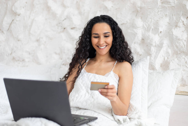 Ευτυχισμένος νεαρός καυκάσιος μελαχρινή κυρία με μακριά σγουρά μαλλιά σε πιτζάμες κάνουν online ψώνια με πιστωτική κάρτα και φορητό υπολογιστή - Φωτογραφία, εικόνα