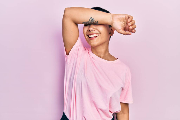 Piękna Latynoska kobieta z krótkimi włosami w luźnej różowej koszuli pokrywającej oczy uśmiechniętym ramieniem wesołym i zabawnym. ślepa koncepcja.  - Zdjęcie, obraz