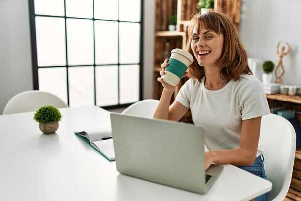 Genç beyaz kadın dizüstü bilgisayar kullanıyor. Kahve içiyor. Evdeki masada oturuyor. - Fotoğraf, Görsel
