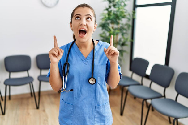 Junge brünette Ärztin im Wartezimmer erstaunt und überrascht, wie sie mit erhobenen Armen und erhobenen Fingern nach oben schaut und zeigt.  - Foto, Bild