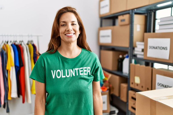 Eine hispanische Frau mittleren Alters, die am Spendenstand ein freiwilliges T-Shirt trägt, sieht positiv und glücklich aus und lächelt mit einem selbstbewussten Lächeln, das Zähne zeigt  - Foto, Bild