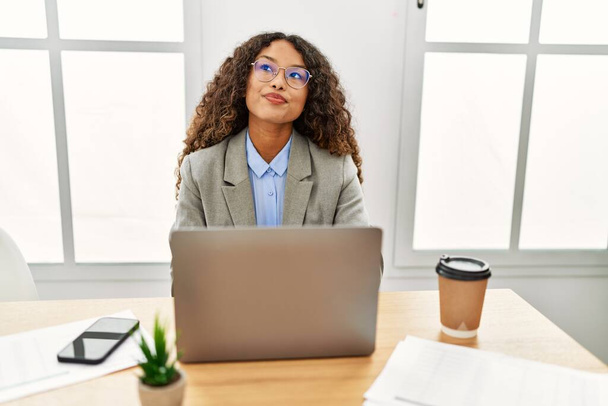 Piękna latynoska kobieta biznesu siedzi na biurku w biurze i pracuje z laptopem uśmiechnięta patrząc w bok i patrząc myśląc.  - Zdjęcie, obraz