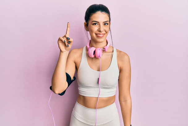 Mujer hispana joven que usa ropa de gimnasio y usa auriculares que muestran y señalan con el dedo número uno mientras sonríe confiada y feliz.  - Foto, imagen