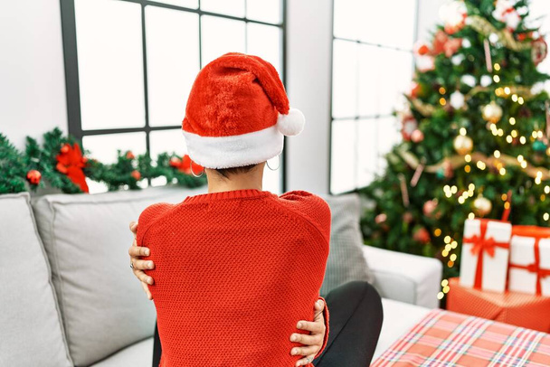 Young hispanic woman with short hair wearing christmas hat sitting on the sofa hugging oneself happy and positive from backwards. self love and self care  - 写真・画像