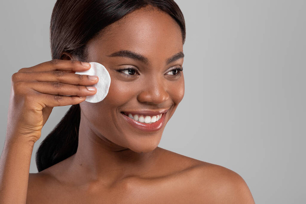 Happy pretty black woman with perfect skin removing makeup with cotton pad isolated on gray background - 写真・画像