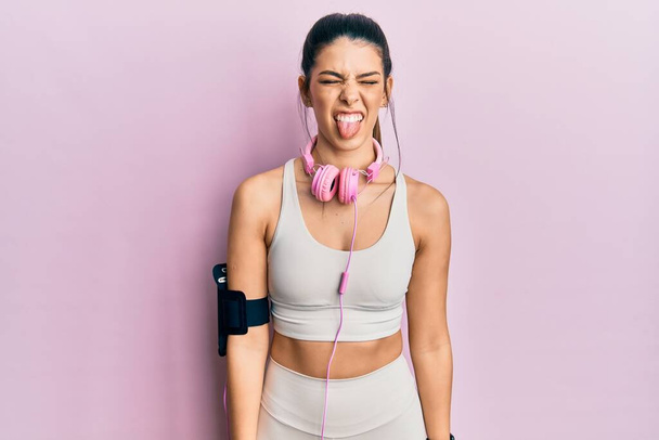 Jonge Spaanse vrouw draagt sportkleding en gebruikt een koptelefoon die haar tong uitsteekt, blij met een grappige uitdrukking. emotie concept.  - Foto, afbeelding