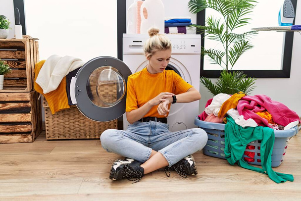 Nuori blondi nainen tekee pyykkiä istuu pesukone tarkistaa aikaa rannekellon, rento ja luottavainen  - Valokuva, kuva