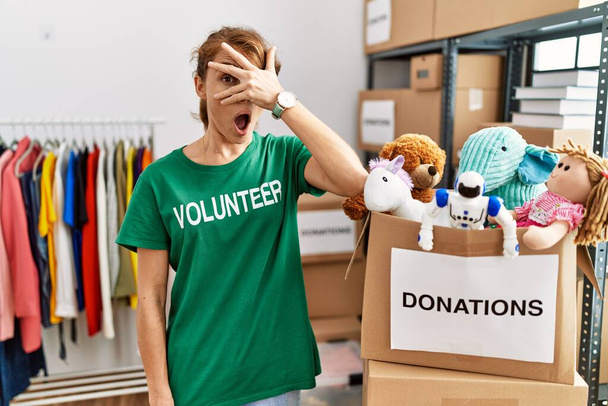 Beautiful caucasian woman wearing volunteer t shirt at donations stand peeking in shock covering face and eyes with hand, looking through fingers with embarrassed expression.  - Фото, изображение