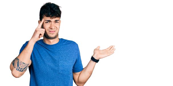 Jovem hispânico vestindo camiseta casual confusa e irritada com a palma aberta mostrando espaço de cópia e apontando o dedo para a testa. Pense nisso..  - Foto, Imagem