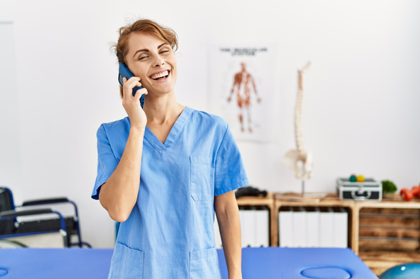 Νεαρή καυκάσια φυσιοθεραπεύτρια χαμογελά ευτυχισμένη μιλώντας στο smartphone της κλινικής. - Φωτογραφία, εικόνα