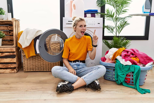 Young blonde woman doing laundry sitting by washing machine pointing finger up with successful idea. exited and happy. number one.  - Foto, Imagem