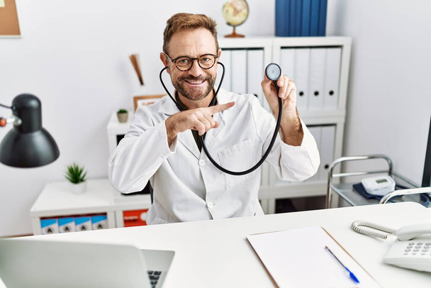Uomo di mezza età con barba indossando uniforme medico tenuta stetoscopio sorridente felice indicando con mano e dito  - Foto, immagini