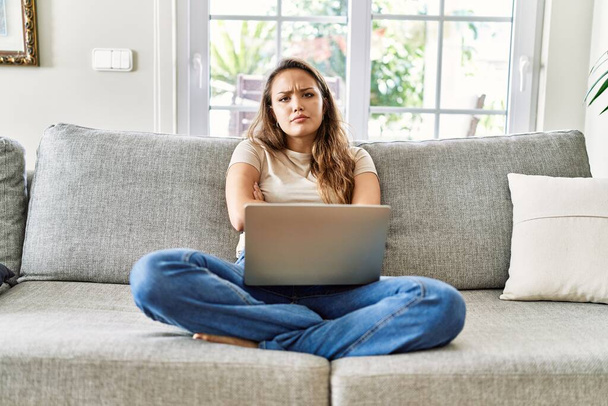 Piękna młoda brunetka siedząca na kanapie, używająca laptopa komputerowego w domu, sceptyczna i nerwowa, dezawująca wyraz twarzy skrzyżowanymi ramionami. osoba negatywna.  - Zdjęcie, obraz