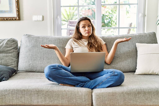 Όμορφη νεαρή μελαχρινή γυναίκα κάθεται στον καναπέ χρησιμοποιώντας φορητό υπολογιστή στο σπίτι ανίδεη και συγκεχυμένη έκφραση με τα χέρια και τα χέρια ψηλά. έννοια της αμφιβολίας.  - Φωτογραφία, εικόνα