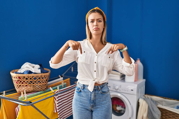 Giovane donna bionda in lavanderia che punta verso il basso guardando triste e sconvolto, indicando la direzione con le dita, infelice e depresso.  - Foto, immagini