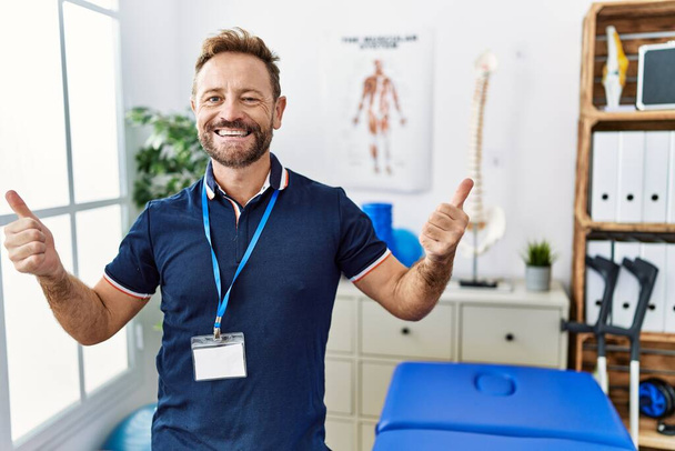 Middle age physiotherapist man working at pain recovery clinic success sign doing positive gesture with hand, thumbs up smiling and happy. cheerful expression and winner gesture.  - Photo, Image