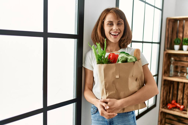 Young caucasian woman smiling confident holding groceries paper bag at home - Foto, imagen