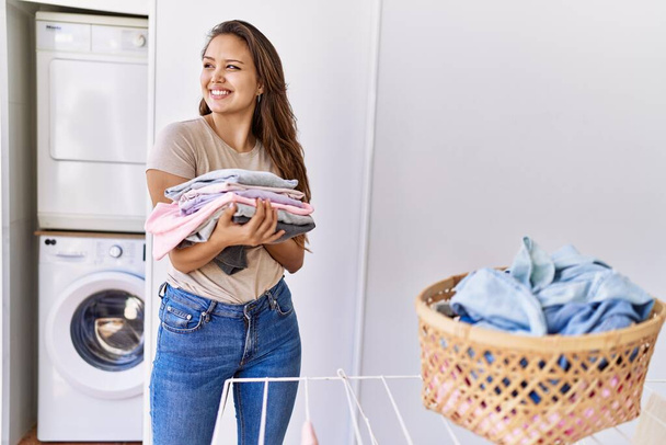 Νεαρή Ισπανίδα κάνει μπουγάδα κρατώντας διπλωμένα ρούχα στο σπίτι. - Φωτογραφία, εικόνα